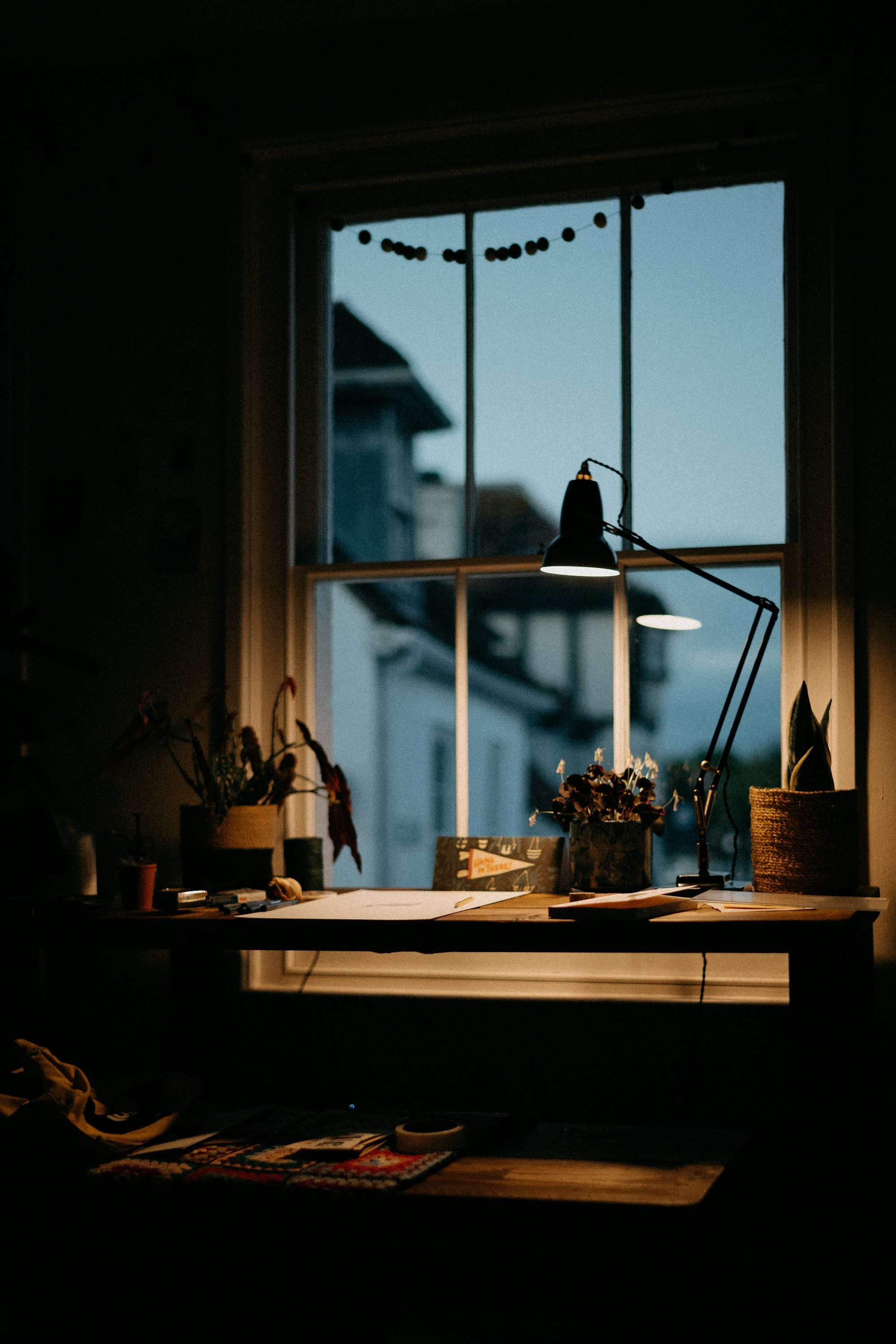Bureau devant une fenêtre à guillotine au crépuscule, une lampe articulée allumée, des plantes.